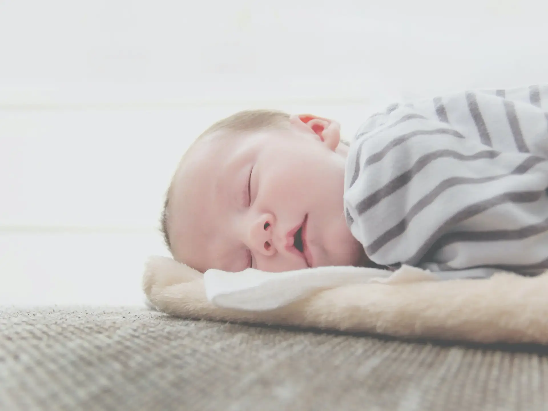 Sleeping newborn resting on a light carpet with a gray striped blanket partially covering them