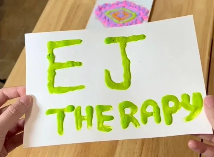 White card with bright lime-green slime letters spelling'EJ THERAPY' (EJ on top, 'THERAPY' beneath), held by two hands.