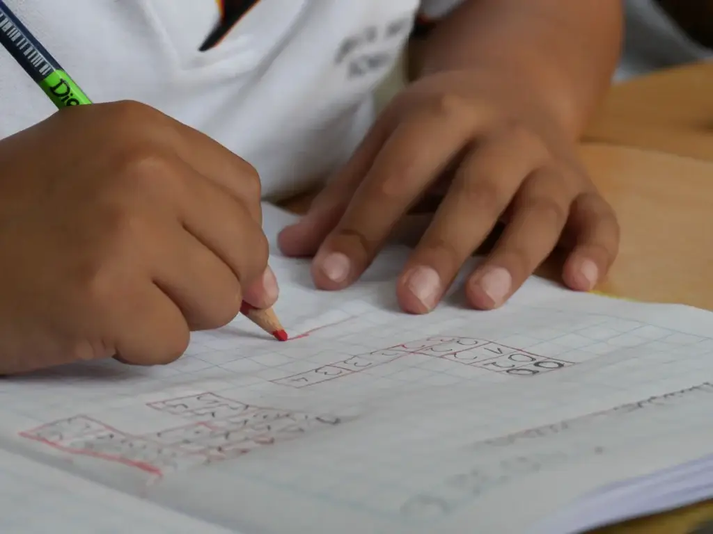 Close-up of a student’s hands writing with a red pencil on graph paper at a desk