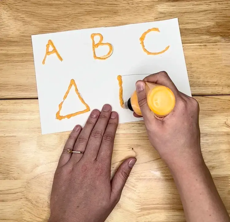 Hands guide an orange glue stick to form the letters A, B, C and a triangle on white paper.