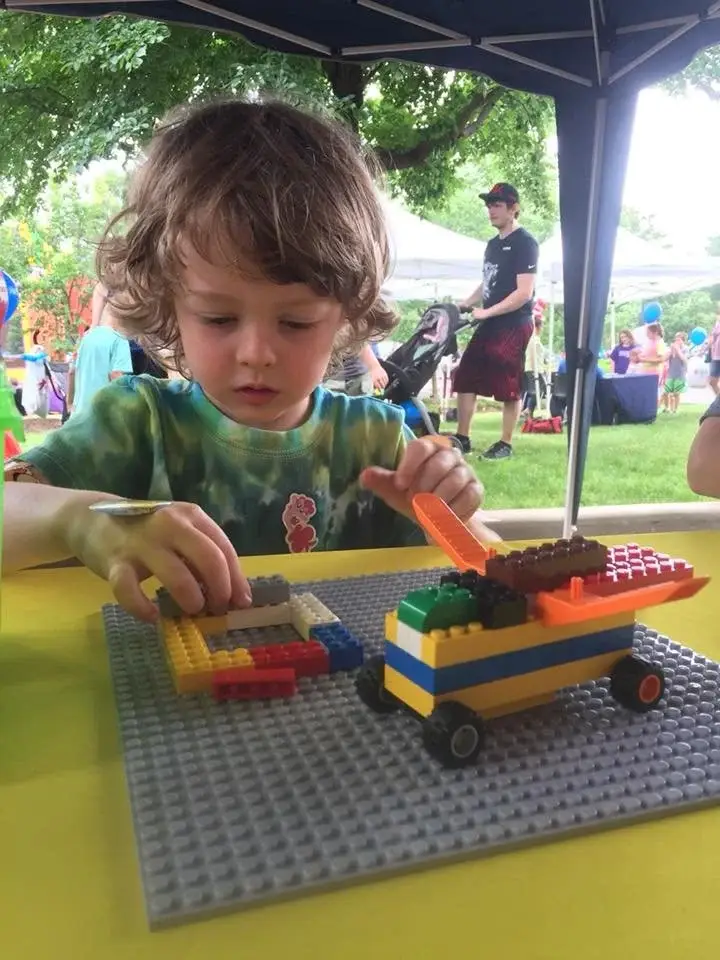 Young child with brown hair builds a colorful LEGO vehicle on a gray baseplate at an outdoor event under a canopy, with people in the background.
