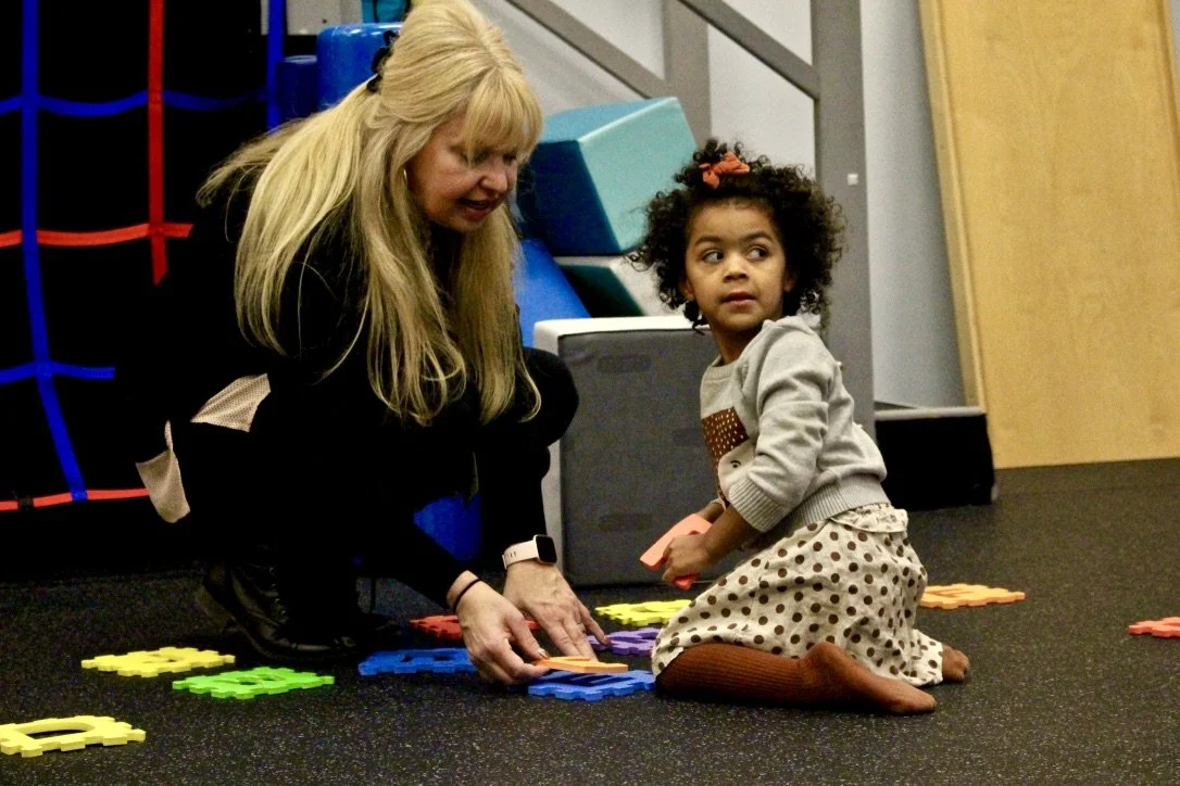 Adult kneels on the floor with a young girl, arranging colorful shape pieces during playtime.