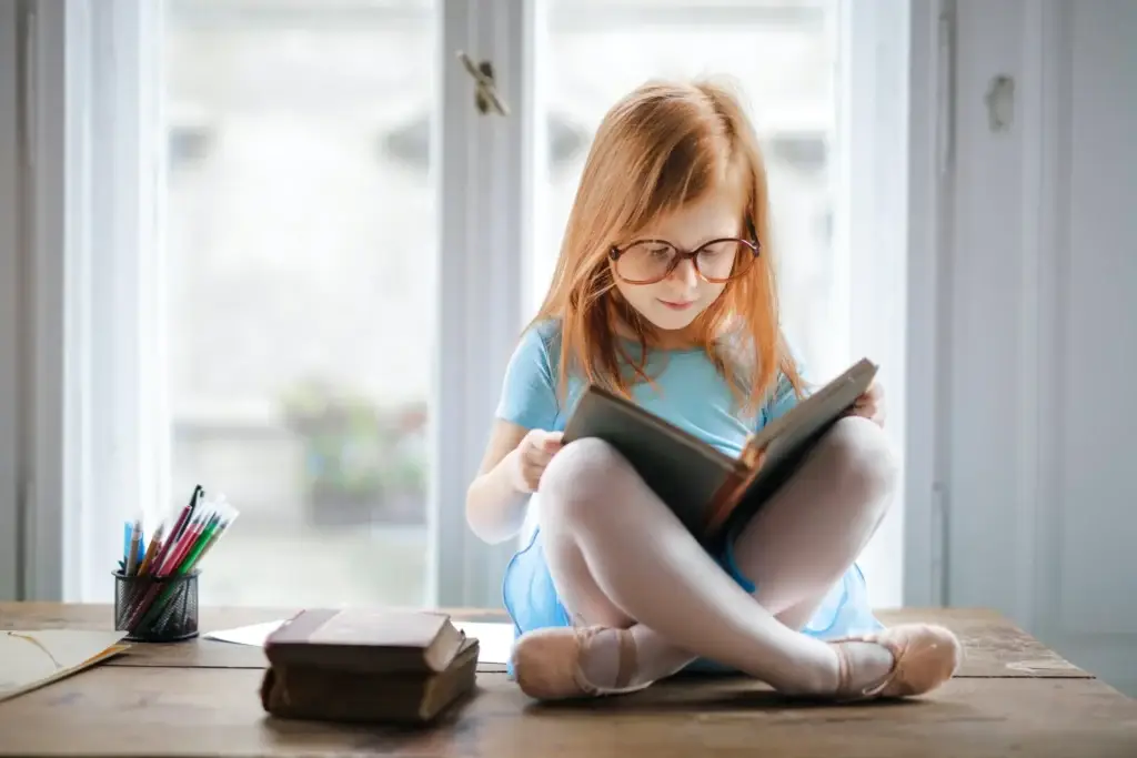 Young girl with glasses reading a book at a wooden table by a sunlit window, pencils in a cup nearby.