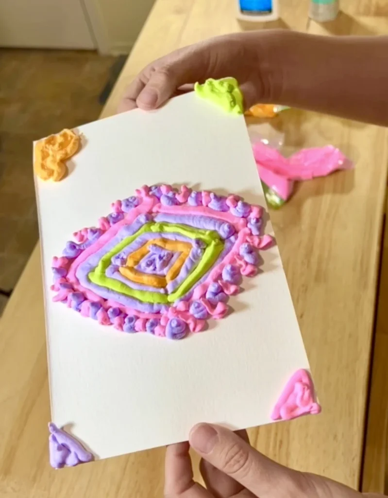 Person holds a white card decorated with a multicolored piping design in a square spiral pattern.