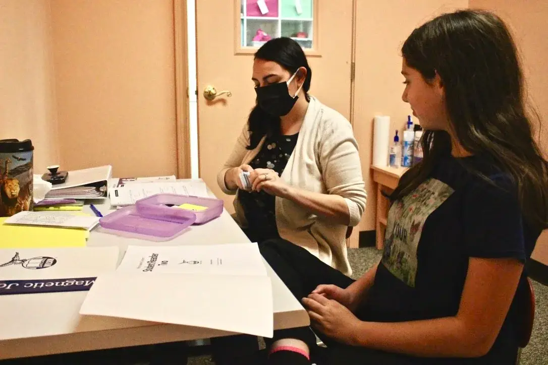 Medical worker wearing a mask and beige cardigan sits at a table with paperwork, handling a small container while a young person sits opposite in a clinic room.