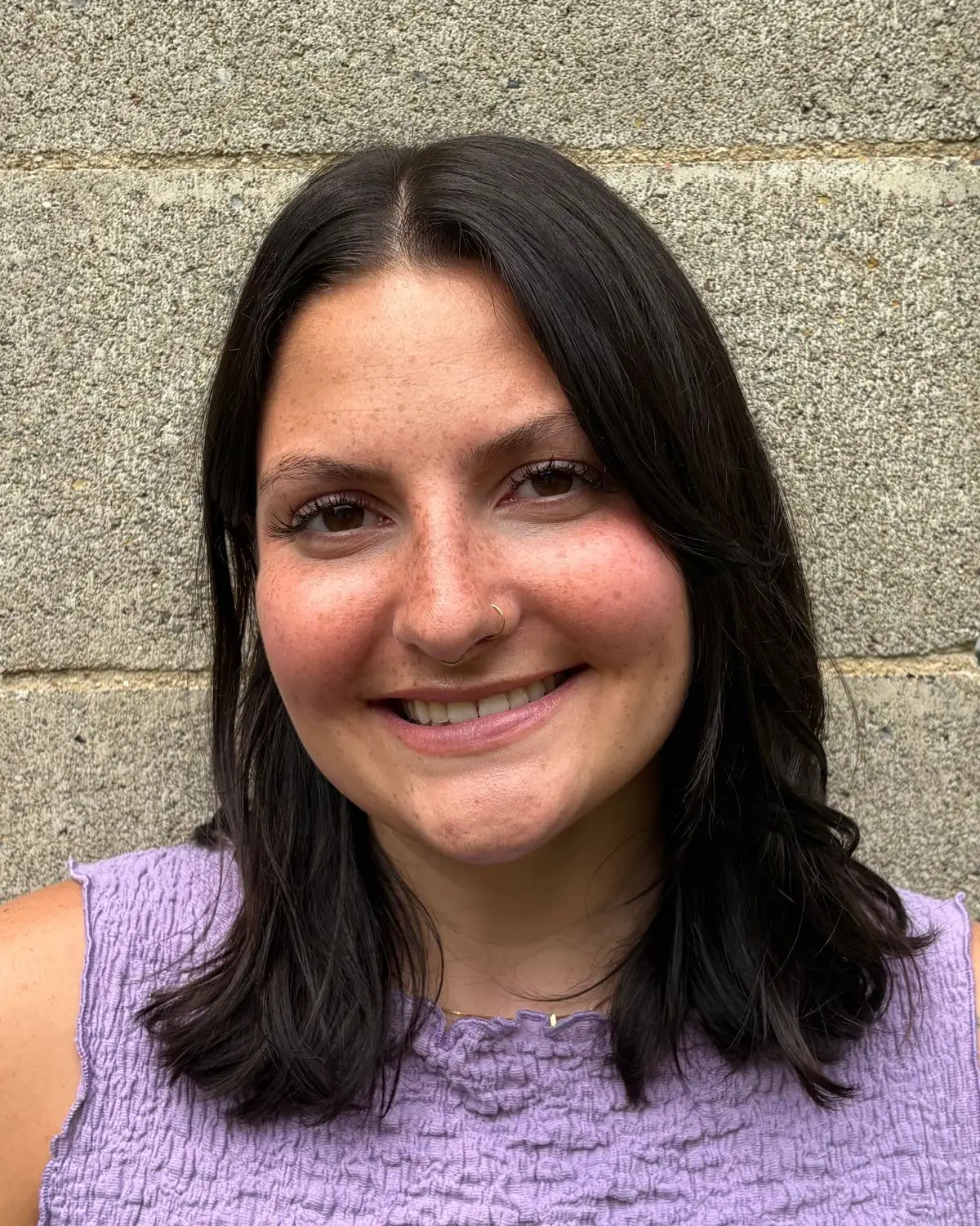 Smiling woman with dark hair and a nose ring, wearing a lavender top, standing against a concrete wall.
