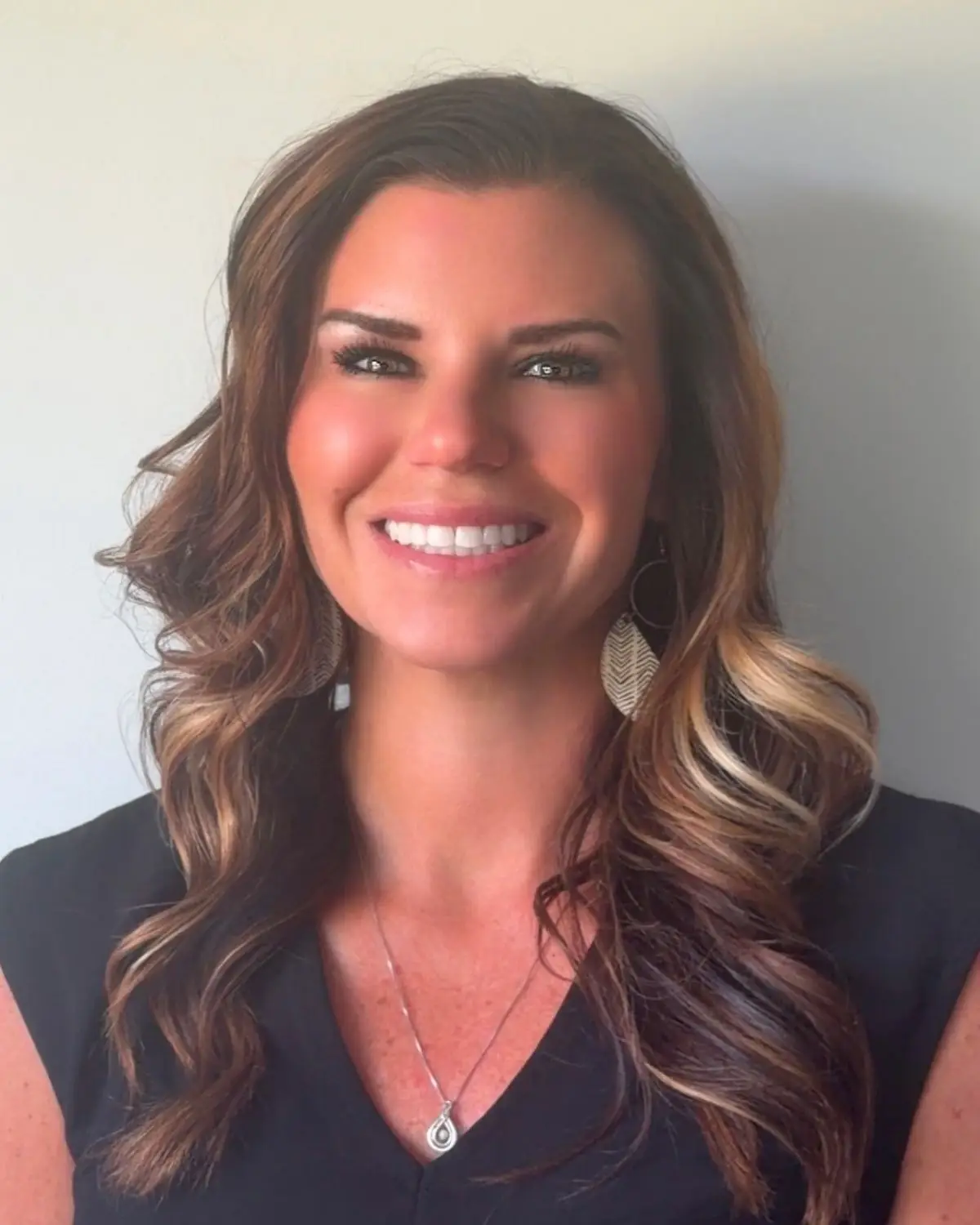 Smiling woman with wavy brown hair, wearing a black blouse, teardrop earrings, and a pendant necklace.