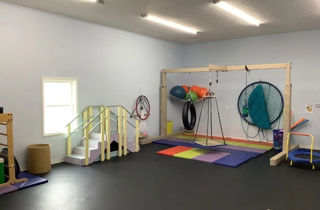 Indoor pediatric therapy gym with colorful mats, swings, rings, and large hoop wall rack for sensory play and obstacle activities.