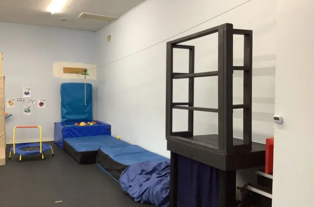 Indoor playroom with blue padded mats, a small trampoline, and a ball pit against a pale wall.