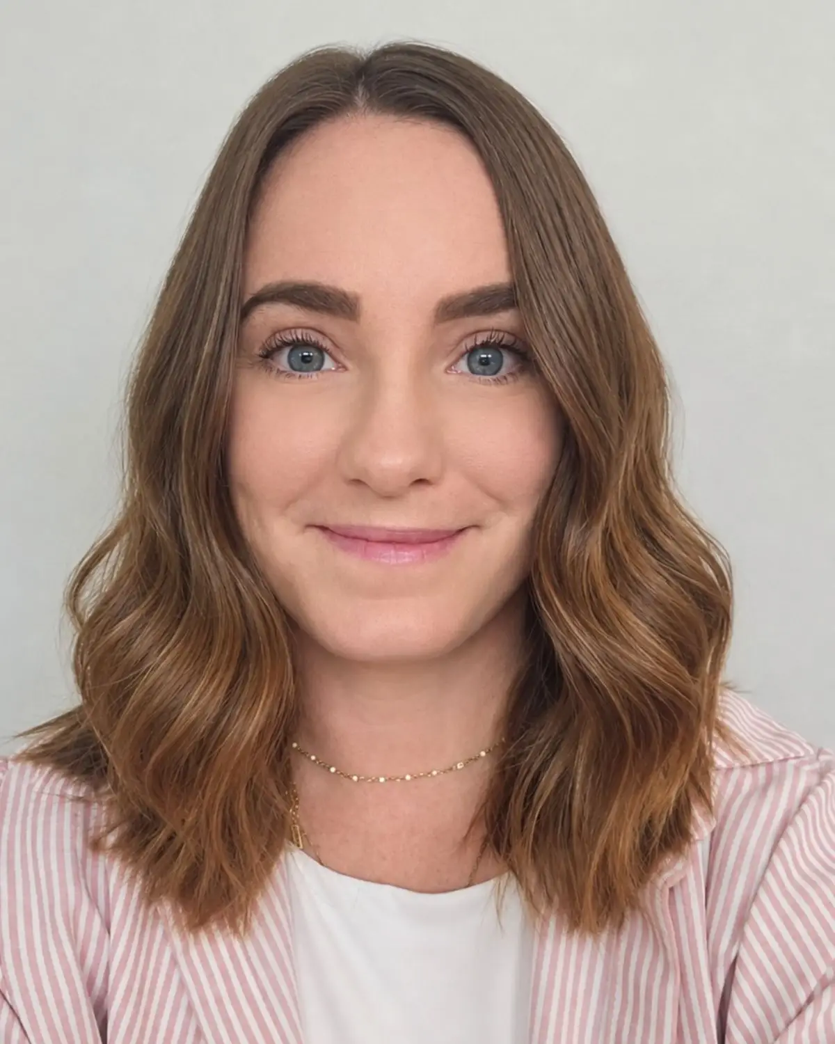 Smiling young woman with shoulder-length, wavy brown hair. Wearing a white shirt, pink, striped blazer, and a necklace against a neutral cream background.