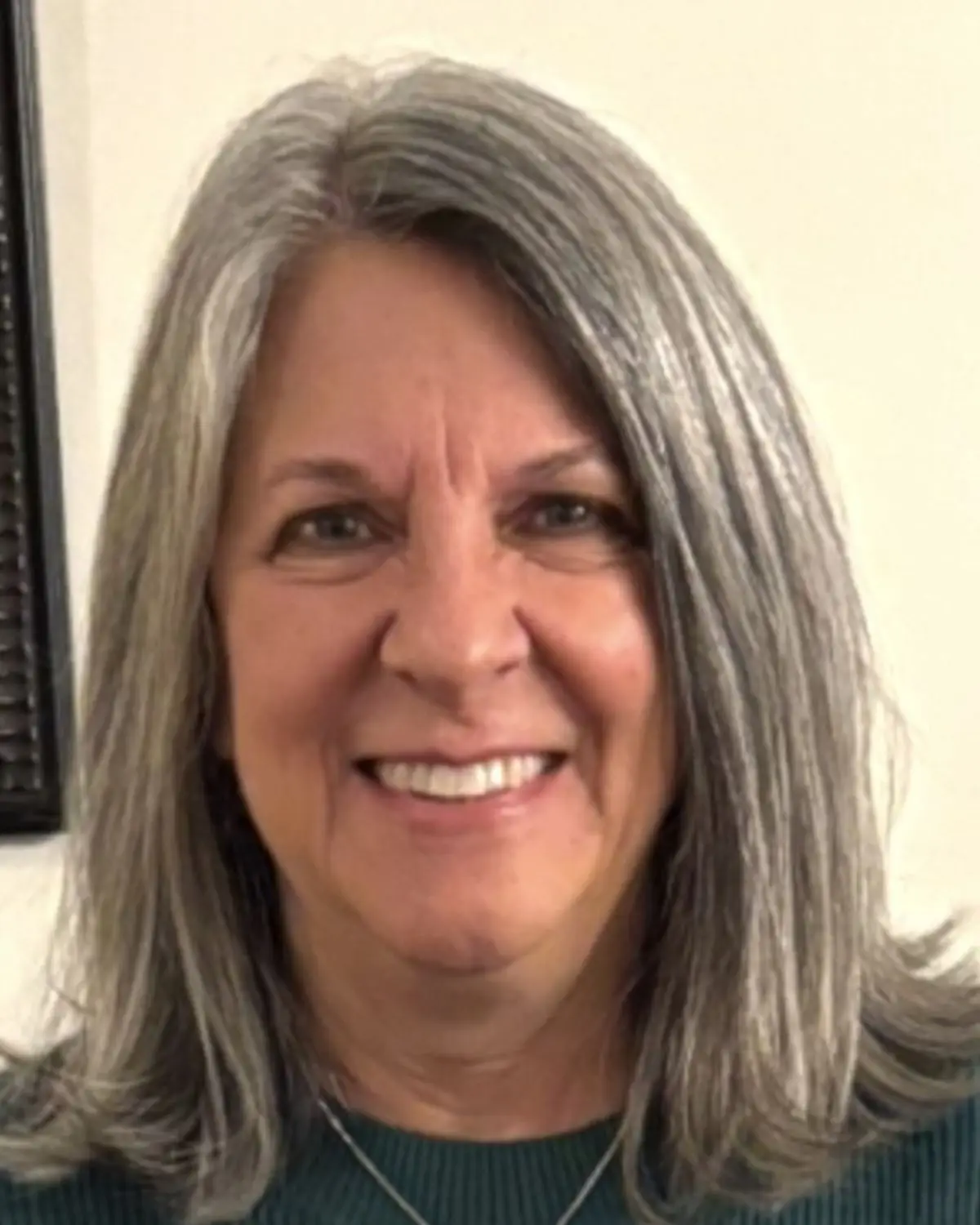Smiling woman with shoulder-length, gray hair wearing a dark green shirt with a necklace, against a yellowish-cream background.