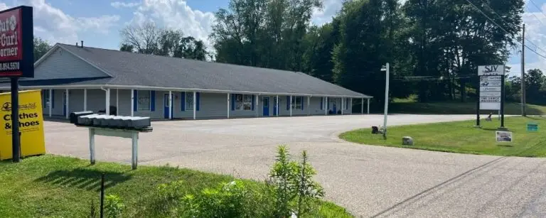 Single-story strip mall with blue doors along a long storefront, a gravel parking lot, and green grass on a sunny day.