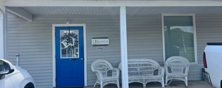 Front porch of a house with a bright blue door, white wicker seating, and a wall sign that appears to say 'Pet Therapy'.