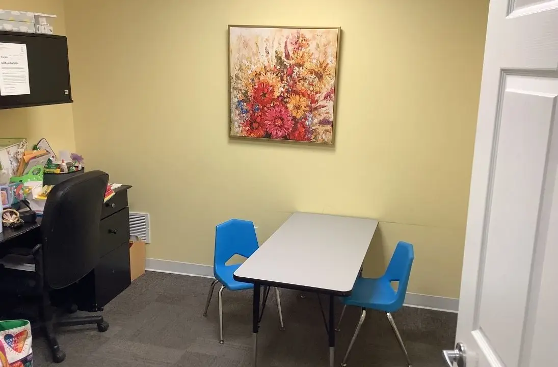 Small office corner with a desk, chair, and filing cabinet; a light gray table with two blue child-sized chairs against a pale yellow wall, and a colorful flower painting above.