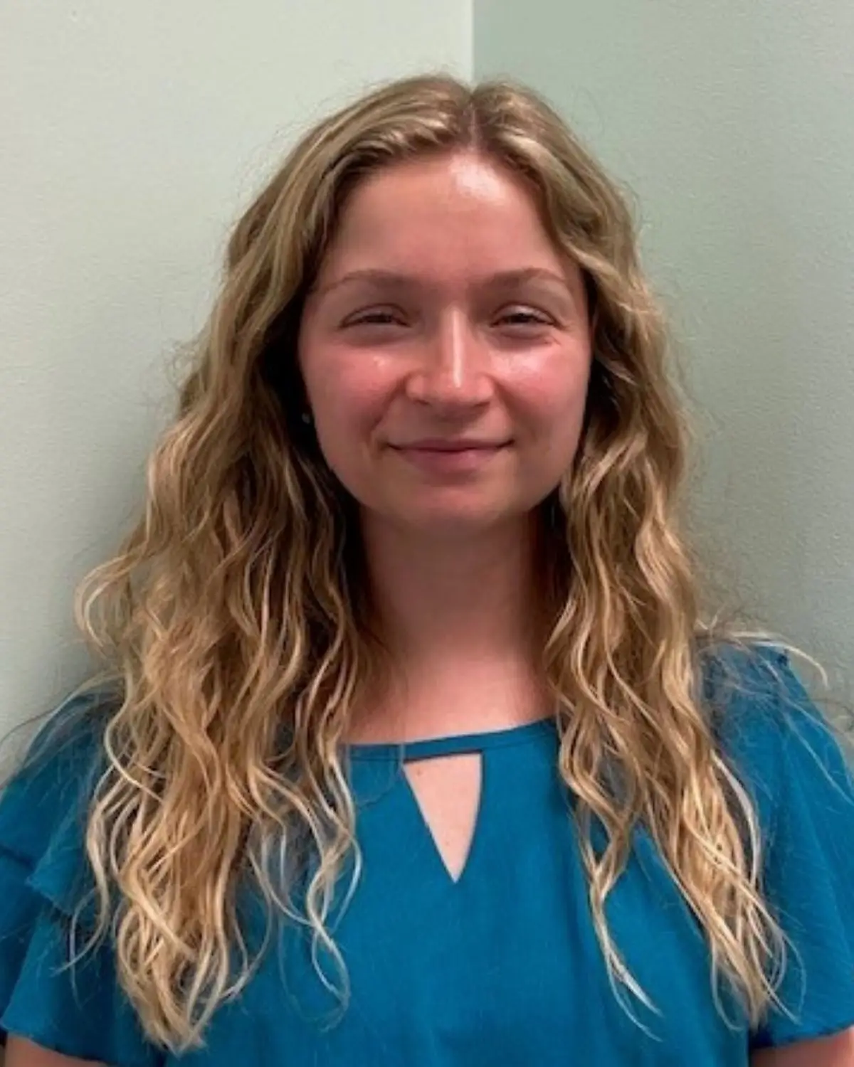 Young, smiling woman wearing a blue t-shirt with long, wavy, blonde hair. Pictured against a greenish gray background.
