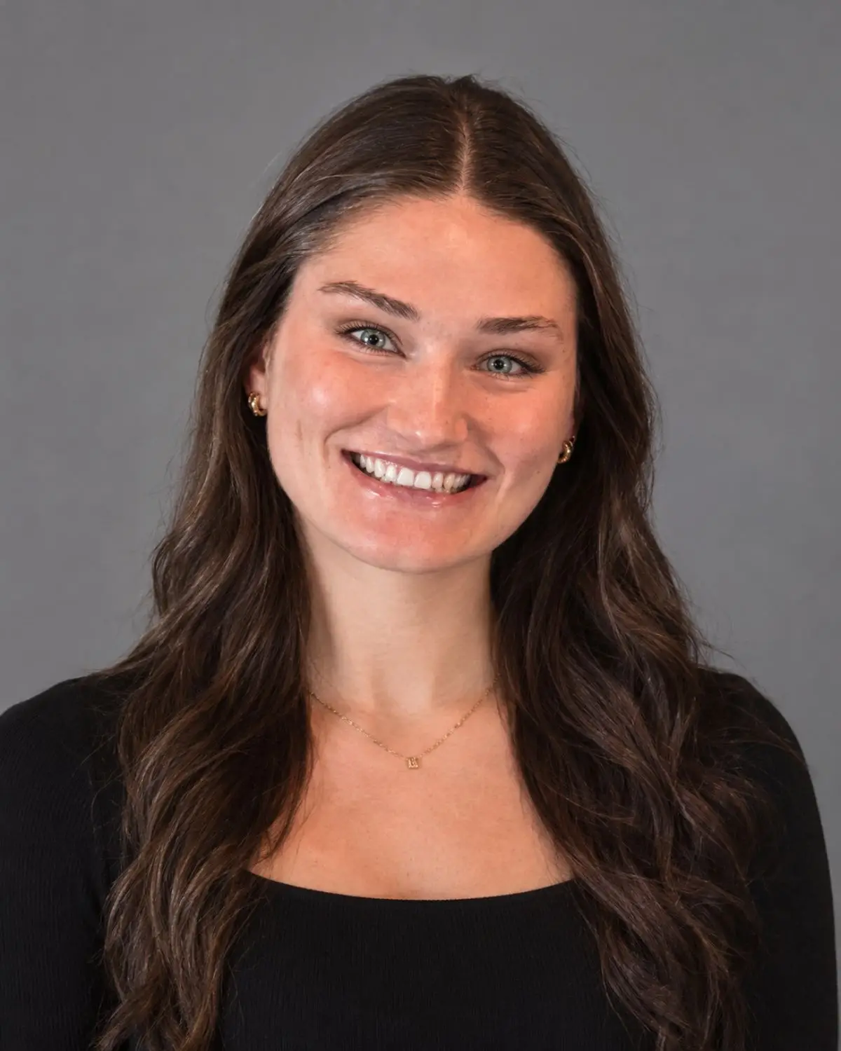 Young, smiling woman with long, wavy, dark brown hair. She is wearing a necklace, small hoop earrings, and a black shirt, and the picture is set against a gray background.