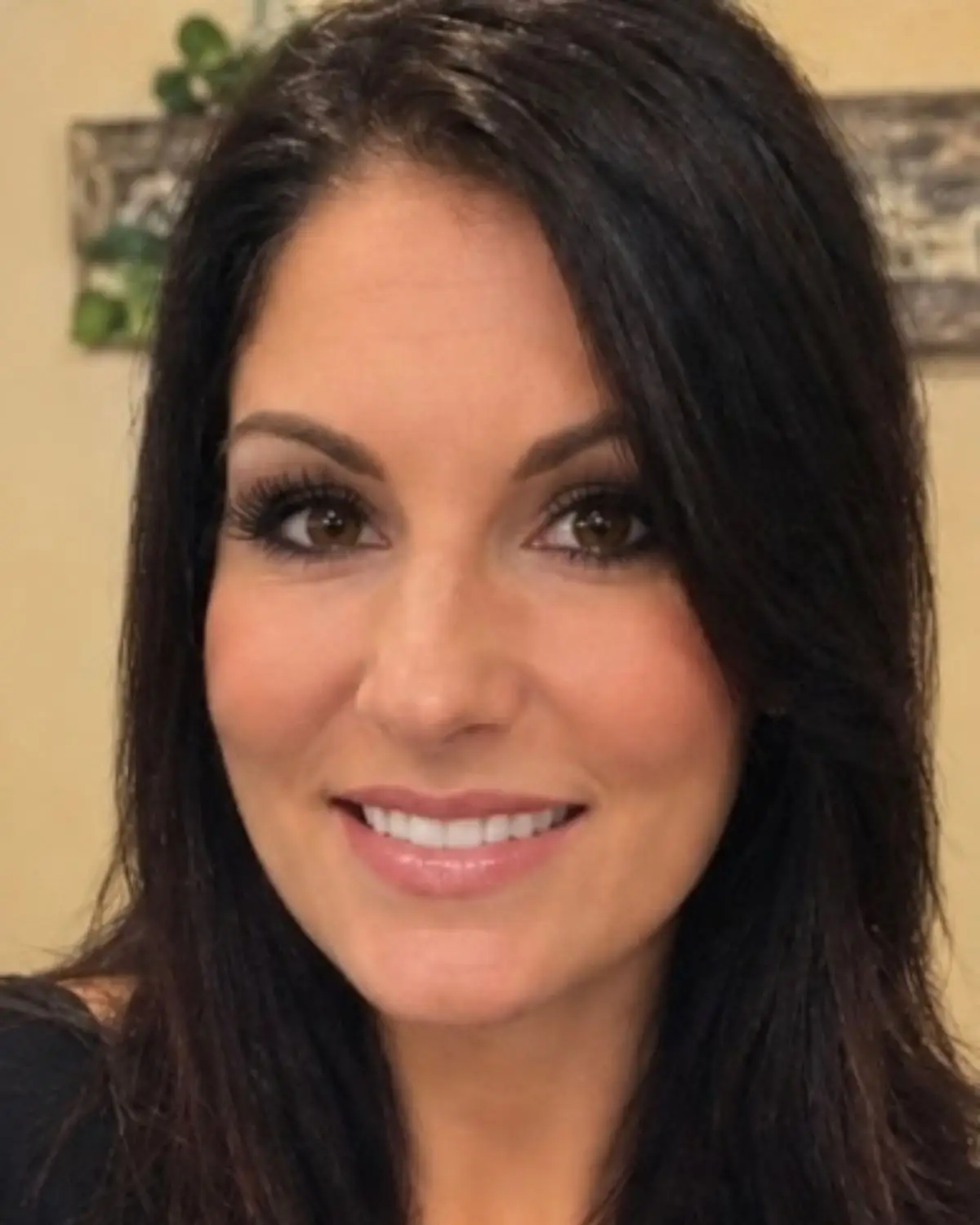 Smiling woman with black hair, smoky eye makeup look, and wearing a black shirt. The picture is set against a yellow background.